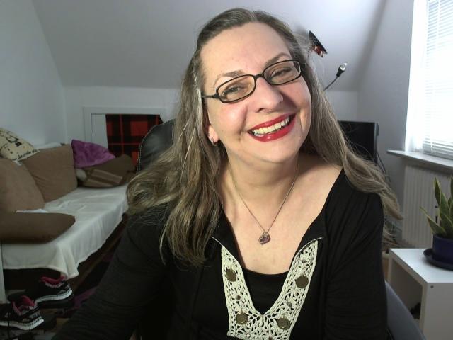 CashLadyVivian: bella, con occhiali, rossetto rosso, top nero, bordi in pizzo bianco, sorridente, capelli castani, collana, seduta in una stanza, pelle chiara, carina, attraente..