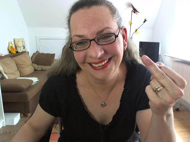 CashLadyVivian – heiß, mit Brille, rotem Lippenstift und schwarzem Oberteil, lächelnd, hält ihren Zeigefinger mit einem goldenen Ring hoch und zeigt ihre Brüste subtil..