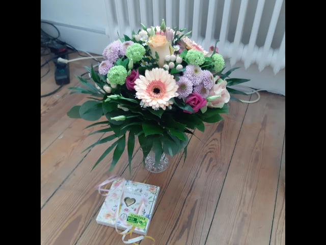 Un ramo de flores con gerberas rosas y blancas, crisantemos morados y verdes, y rosas rosadas en un jarrón de vidrio sobre un suelo de madera, con una tarjeta..