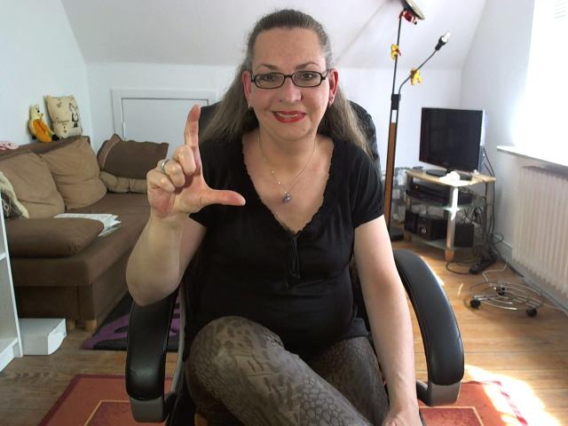 CashLadyVivian-hot, uma mulher rechonchuda com óculos, blusa preta e calças de legging com estampa de leopardo, está sentada em uma cadeira, apontando com o dedo indicador e sorrindo. Suas nádegas estão ligeiramente visíveis..