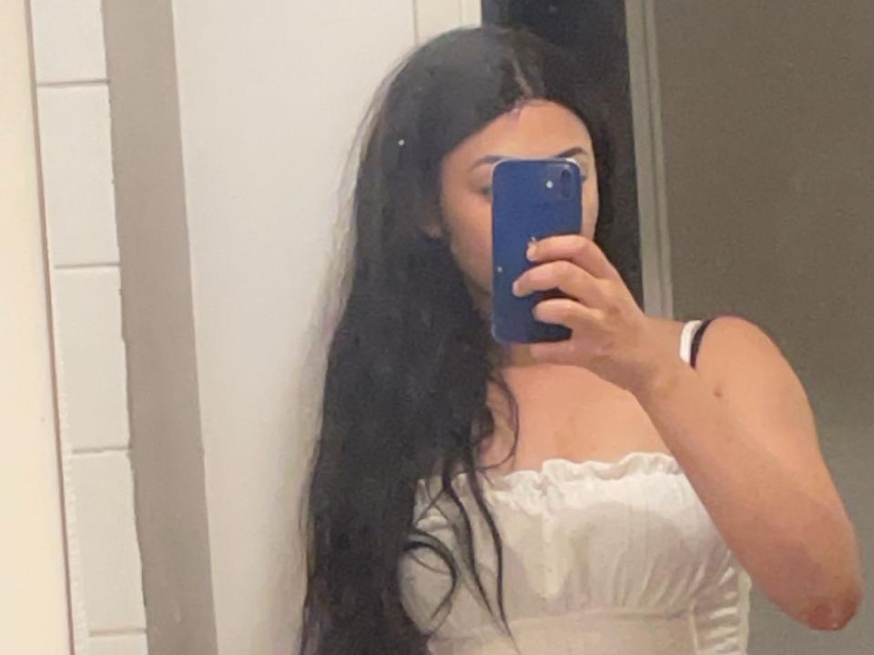LeaSeven with long black hair, holding a blue phone, wearing a white strapless top, showing her boobs in a mirror selfie.