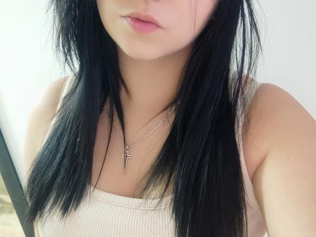 Kkatalina has black, straight hair, pink lips, and a light skin tone. She's wearing a beige tank top with a silver cross necklace. Her small tits are visible.