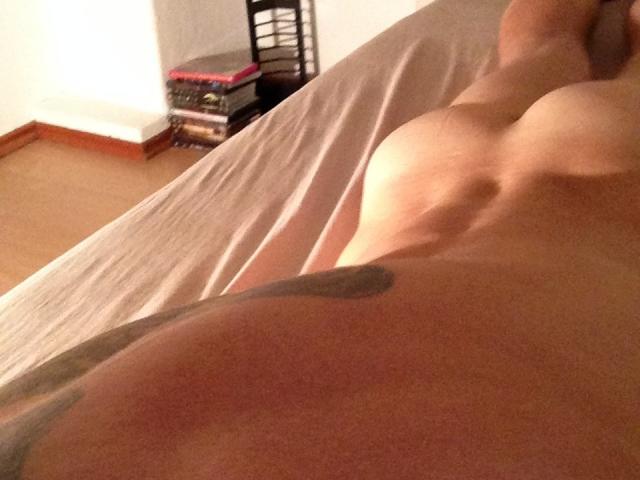 Surviver's naked upper body, showing small tits and a tattoo on the side, lying on a bed with a beige sheet. Survivor's legs are partially visible.