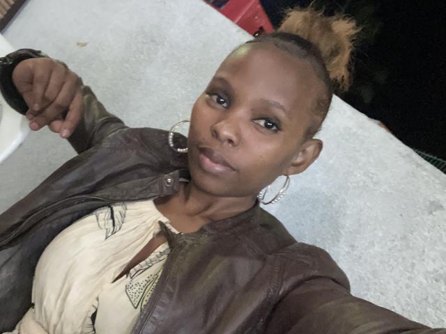 CynthiaAnx with dark skin, small hoop earrings, and a brown leather jacket, wearing a beige top with a patterned neckline, her hair in a top knot, and a relaxed expression.
