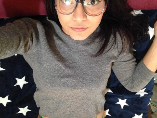 AriaLeenn with glasses, grey t-shirt, lying on a star-patterned blanket, looking up at the camera, medium-length dark hair, slight smile, visible bare pussy at the bottom.