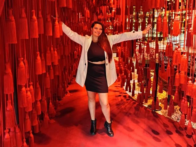 KatherinLatin está de braços abertos, vestindo um casaco branco sobre uma cropped preta e uma minissaia, botas pretas, e sorrindo. Decorações com franjas vermelhas estão penduradas ao redor dela..