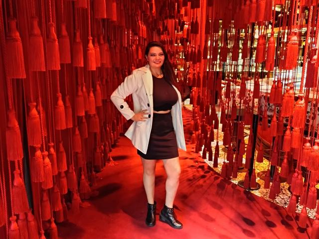 KatherinLatin with dark hair, white jacket, black dress, belt, black boots, smiling, standing in red tassel-filled hallway. Boobs visible under dress, ass covered by dress.