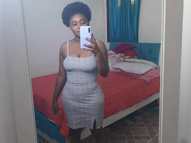 EbonyKath vestindo um vestido cinza justo com uma fenda, mostrando suas curvas e seios pequenos, tirando uma selfie no espelho com um celular branco em um quarto..