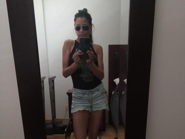 LorenaM has tanned skin, dark hair in a bun, wearing sunglasses, a black tank top, and frayed denim shorts. She's holding a phone, standing in front of a mirror.