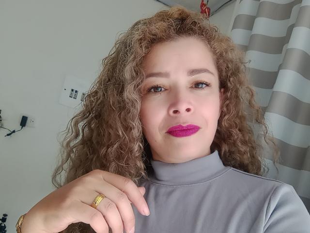 A DalilaSuperHot tem cabelo castanho encaracolado, pele clara, batom roxo e um suéter de gola alta cinza. Ela está segurando um pano marrom e tem um anel de ouro no dedo..