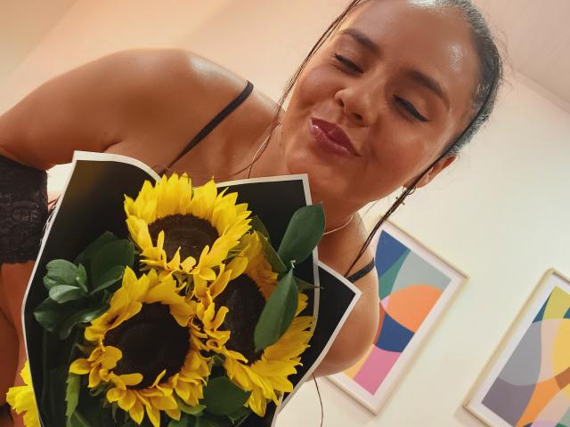ArielleWilde with dark hair in a bun, wearing black lace, holding sunflowers and yellow flowers, smiling, wearing pink lipstick, and earbuds, in a slightly low angle shot.