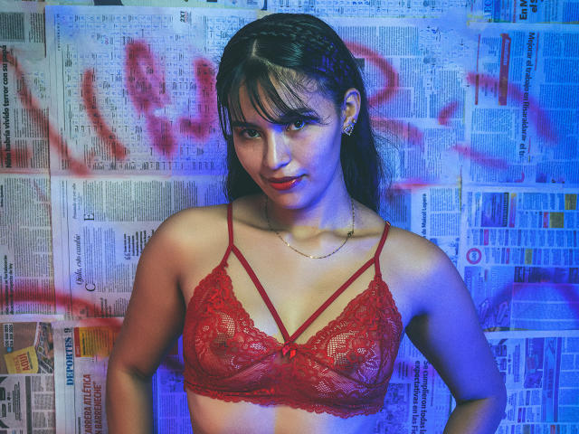 SusanKartier with dark hair, bangs, and red lace bra, showing small tits, standing in front of newspaper wall with red graffiti, looking at camera with slight smile.
