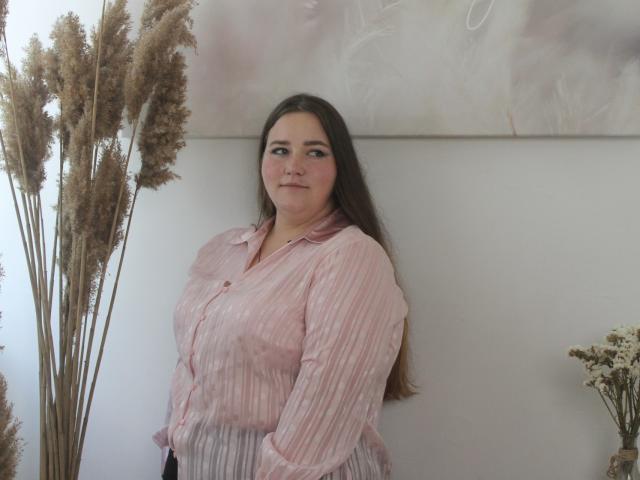 DaniellaGraceZZ, barefoot, sitting on chair, holding white bouquet, pink striped shirt, black pants, long brown hair, smiling, wooden floor, potted plant, wooden cabinet, vase, flowers.