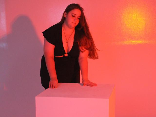 DaniellaGraceZZ with brown hair, pink striped shirt, holding white cup and saucer, slightly smirking, sitting on gray chair, white wall, black framed pictures, and white lamp in background.