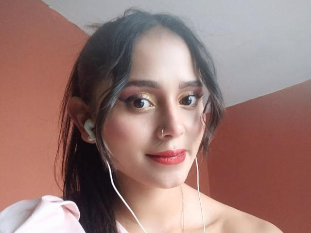 Eli Baker tiene el pelo oscuro, lápiz labial rojo y auriculares blancos puestos, además de un piercing en la nariz y un maquillaje discreto. Está sonriendo ligeramente, mostrando sus labios carnosos. Sus senos no son visibles..