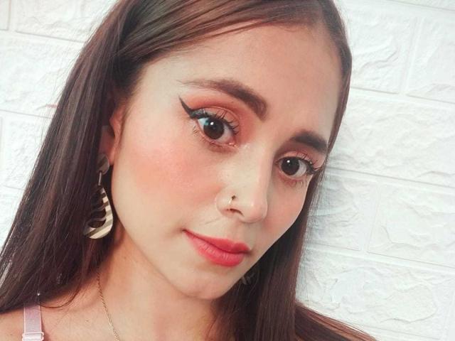 EliBaker a les yeux marron, du rouge à lèvres, un trait d'eye-liner ailé et un piercing au nez. Elle porte des boucles d'oreilles en forme de croissant de lune et a de longs cheveux bruns. Son visage est légèrement rougi..
