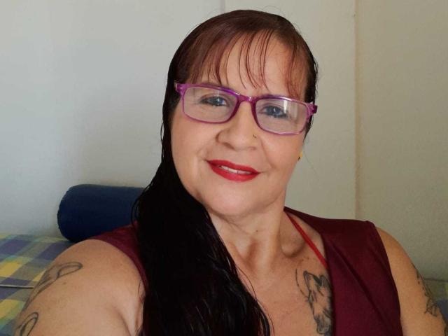 Laura Riley, con lápiz labial rojo, gafas moradas y cabello negro mojado, sonriendo a la cámara. Lleva puesto un top rojo sin mangas, que muestra tatuajes en sus brazos y pecho..
