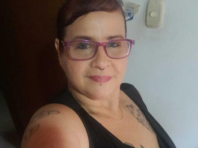 Laura Riley tiene el pelo corto y castaño, gafas moradas, un piercing en la nariz y una camiseta negra sin mangas que muestra tatuajes en los hombros. Sus pechos son pequeños y está sonriendo..