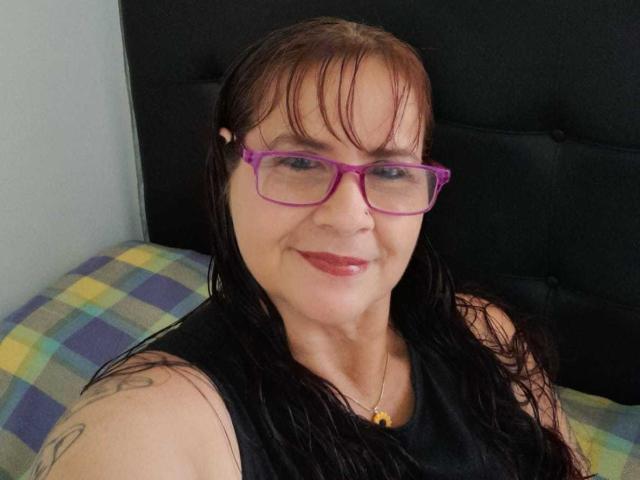 LauraRiley with wet brown hair, pink glasses, black tank top, nose ring, tattoo on shoulder, and red lipstick, smiling at the camera. Her tits are average-sized.