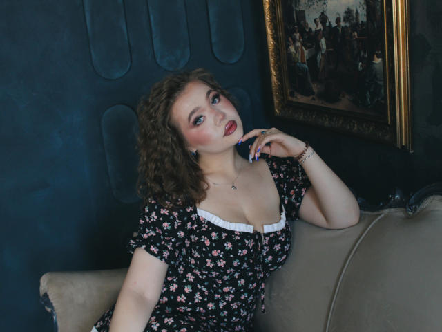 StacyYellig with curly brown hair, wearing a floral dress, leaning on a beige sofa, hand near mouth, red lipstick, blue eyes, white chest trim, subtle cleavage.