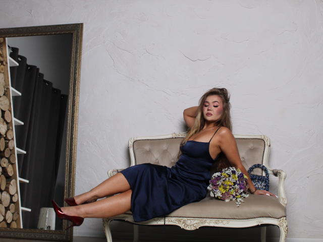 StacyYellig is sitting on a beige, tufted, vintage-style couch wearing a dark blue, strapless dress and red heels, with a bouquet of flowers in her lap, eyes closed.