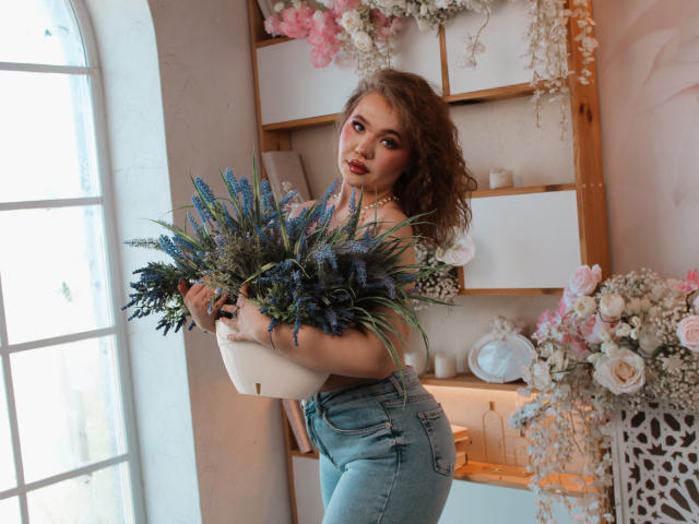 StacyYellig with curly brown hair, holding a bouquet of blue and green flowers, wearing high-waisted blue jeans, and a pearl necklace, standing by a window.