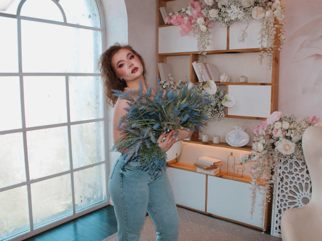 Stacy Yellig, avec ses cheveux bruns bouclés, tenant un bouquet de fleurs bleues et vertes, portant un pantalon en jean taille haute, se tenant près d'une grande fenêtre, une étagère blanche avec des fleurs en arrière-plan..