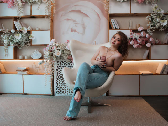 StacyYellig with curly brown hair, topless, holding pink phone, wearing blue jeans, sitting in white chair, holding necklace, barefoot, floral and bookshelf background, soft pink rose wall art.