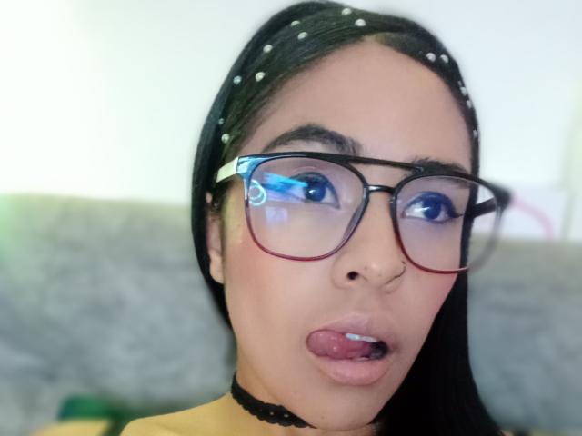 StellaBeckker with black polka-dot headscarf, red-framed glasses, choker, and tongue out, showing her puckered lips and small nose ring. She has long black hair and a light brown skin tone.