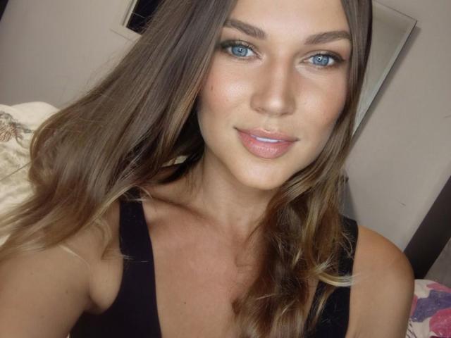BernuLoverx has blue eyes, light brown wavy hair, a cute smile with pink lips, and is wearing a black sleeveless top, showing her bare shoulders and upper chest.