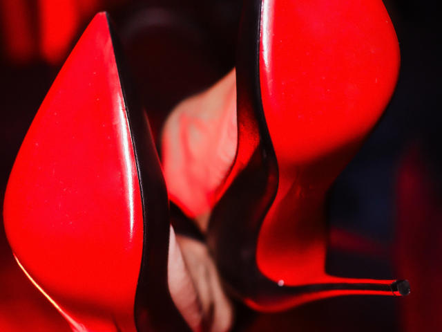 AmberCrost's shiny red stiletto heels, with black soles, are in the foreground. Her toes are visible through the heels, slightly blurred, creating a sexy, high-contrast effect.