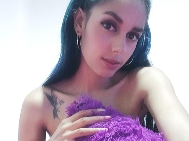 AnaisHilton with blue hair, hoop earrings, and a purple fuzzy pillow, holding it to her bare tits, has a tattoo on her left shoulder, and a subtle smile.