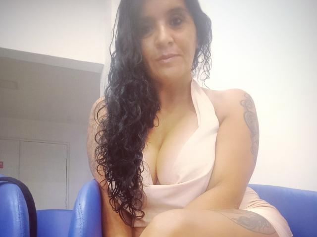 AndysLove with long curly black hair, wearing a pink halter dress, showing big tits and a tattoo on her right arm, sitting on a blue chair.