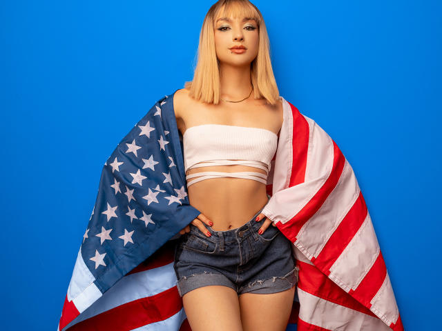 MaryMilan with blonde bob, wearing a white strapless top and denim shorts, draping an American flag over her shoulders, hands on hips, blue background.