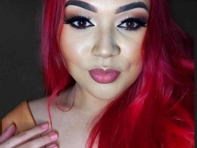GabAmazing has bright red hair, thick black eyebrows, heavy eyeliner, and pink lipstick. She's looking at the camera with a slight smile, touching her hair with one hand.