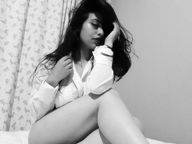 BellaSmithX69 with long dark hair, sitting, wearing an open shirt, exposing her tits, leg raised, hand on her head, black and white, sexy, soft light, gentle expression.