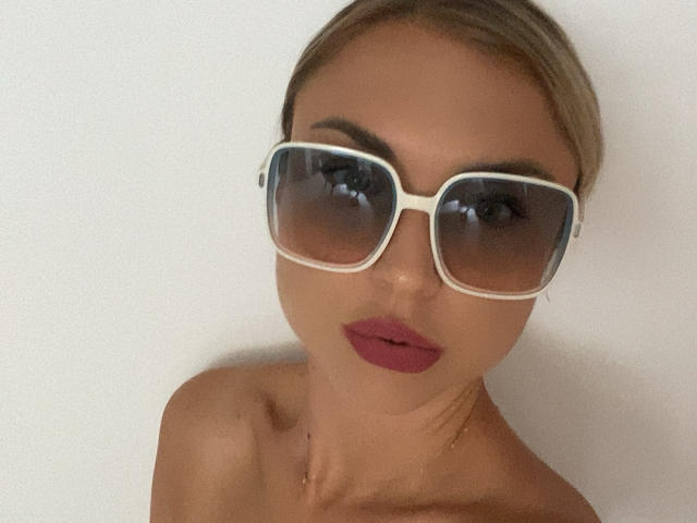 AshleySensual has tan skin, blonde hair, and wears large white sunglasses with dark lenses, and has deep red lipstick on her pouty, nude-boobed face.