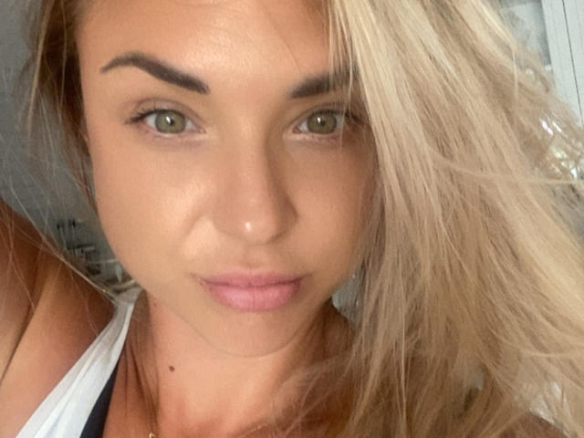 AshleySensual has green eyes, blonde hair, and a small mole under her nose. She's wearing a white tank top, showing her bare shoulders and subtle cleavage.