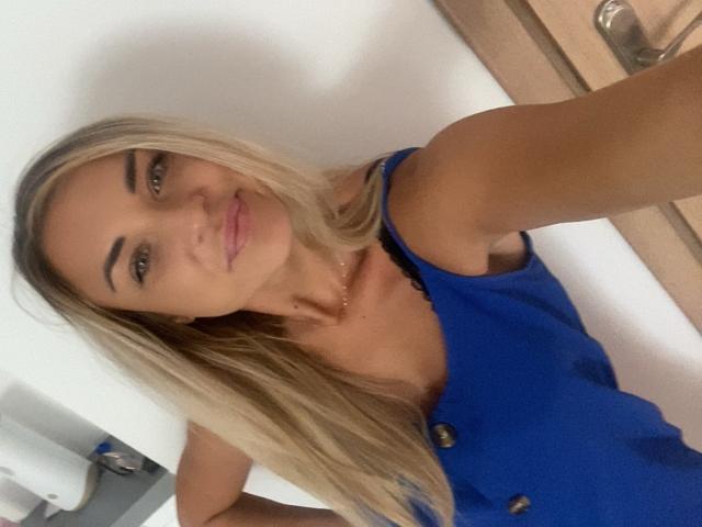 AshleySensual has blonde hair, blue dress with buttons, and is smiling with pink lips. She's taking a selfie, showing her shoulder and upper chest. Her tits are small.