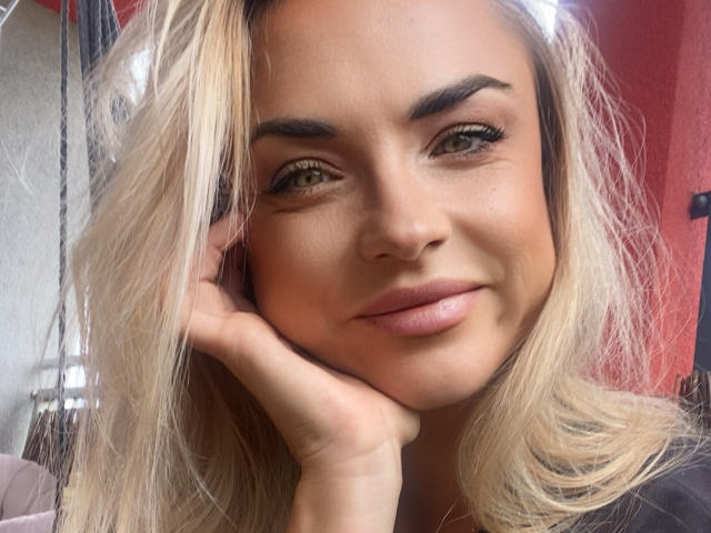 AshleySensual with blonde hair, green eyes, and full pink lips, resting her cheek on her hand, smiling slightly. She has a cute face and is looking directly at the camera.