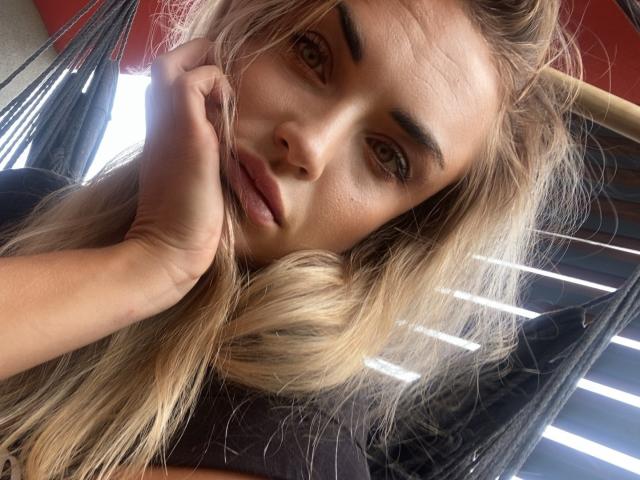 AshleySensual has blonde hair, brown eyes, and full lips, looking seductively at the camera with her hand near her face, wearing a black top, in a low-light setting.