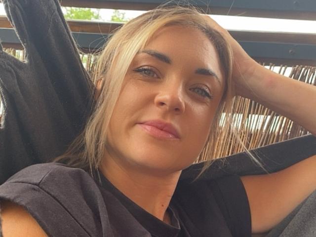AshleySensual, blonde, with light skin, blue eyes, and full pink lips, wearing a black shirt, has a relaxed, slightly smug expression, and rests her head on her hand.