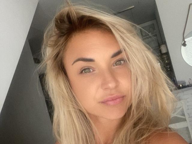AshleySensual has blonde hair, green eyes, and slightly parted pink lips. She's looking at the camera with a subtle smile, her eyebrows are thick and dark.