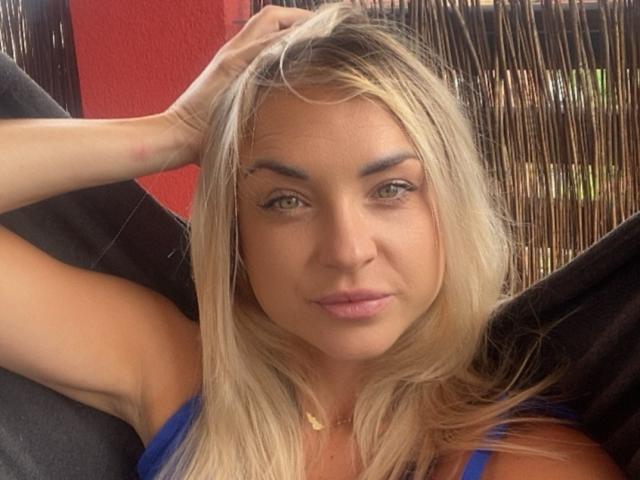 AshleySensual has blonde hair, green eyes, and a slight smile. She's wearing a blue top, with one hand in her hair, and a gold necklace. Her ass is partially visible.