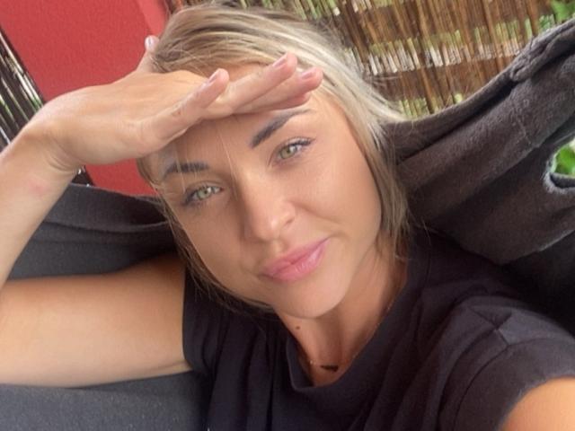 AshleySensual with blonde hair, green eyes, and pink lips, wearing a black shirt, has her hand over her forehead, smiling softly. Her ass is not visible.