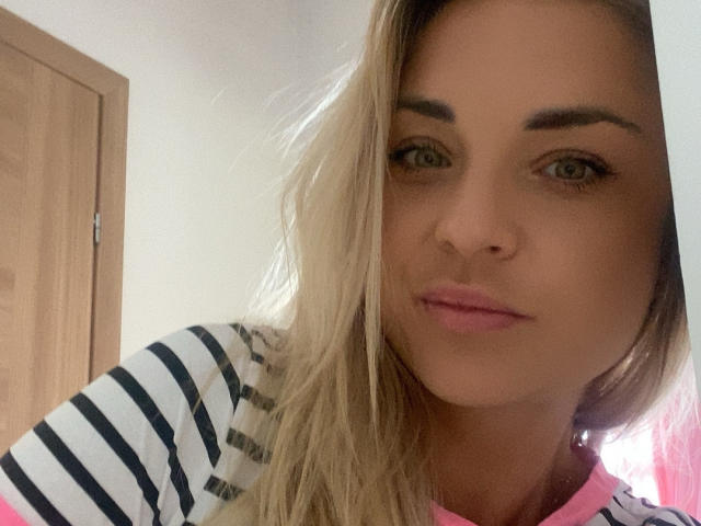 AshleySensual has blonde hair, green eyes, and pink lips. She's wearing a striped shirt with pink trim, looking directly at the camera with a slight pout.