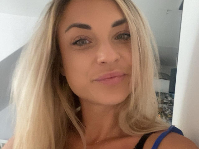AshleySensual has blonde hair, thick eyebrows, green eyes, and pink lips. She's wearing a blue top, showing her small tits, with a slight smile and a cute face.