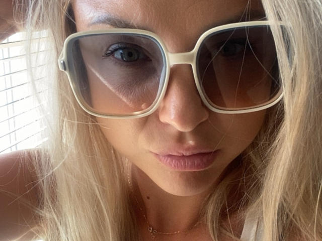 AshleySensual with blonde hair, wearing large beige sunglasses, and a gold necklace, looking directly at the camera with slightly pouty pink lips. Her blue eyes are visible.