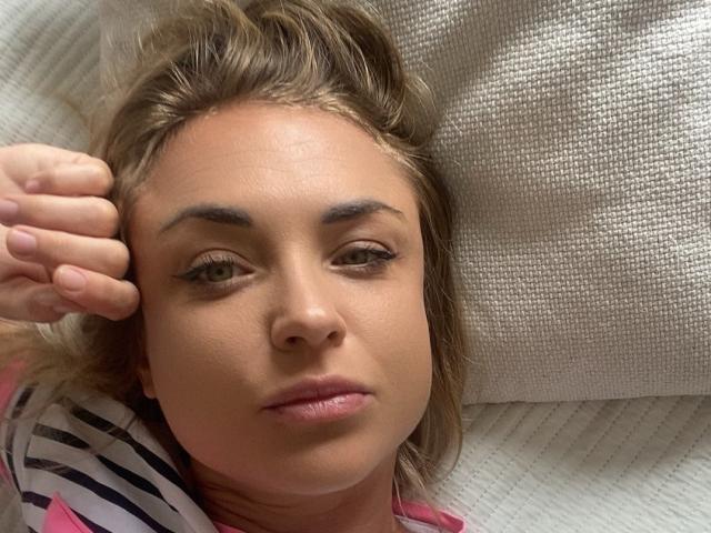 AshleySensual's face, lying down, with blonde hair and brown eyebrows, wearing a pink and black striped shirt, hand on her head, looking slightly tired. Her ass is partially visible.