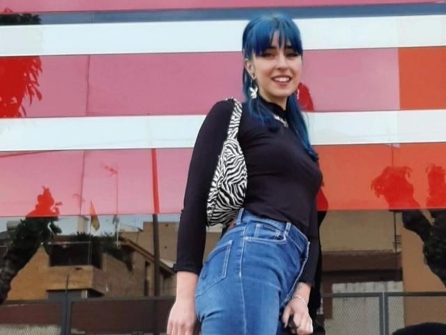 Hanna, com tranças azuis, camisa preta de manga comprida, bolsa com estampa de zebra e jeans azuis, sorrindo, em pé do lado de fora, mostrando o bumbum com os jeans justos..